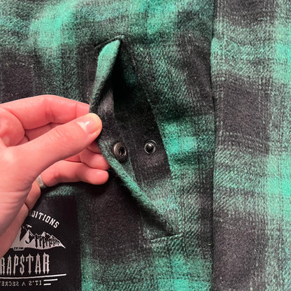 The Trapstar Irongate Green Plaid Lumberjack Jacket is a woven jacket in a green and black checked pattern, made from a 95% polyester and 5% wool outer with a full polyester lining. It features contrast shell panels at the shoulders, exposed popper fastenings at the front, a classic collar, and adjustable popper tabs at the cuffs. Details include side slip pockets, a Trapstar brand patch at the front hem, and a large Trapstar logo embroidered across the upper back yoke.
