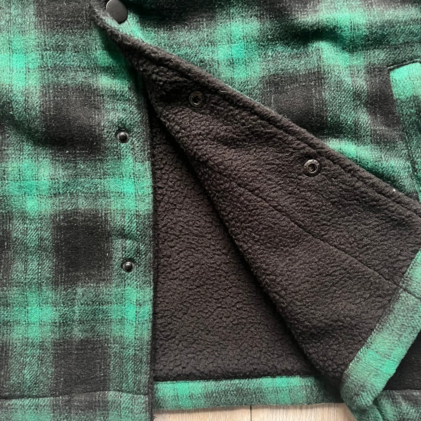 The Trapstar Irongate Green Plaid Lumberjack Jacket is a woven jacket in a green and black checked pattern, made from a 95% polyester and 5% wool outer with a full polyester lining. It features contrast shell panels at the shoulders, exposed popper fastenings at the front, a classic collar, and adjustable popper tabs at the cuffs. Details include side slip pockets, a Trapstar brand patch at the front hem, and a large Trapstar logo embroidered across the upper back yoke.