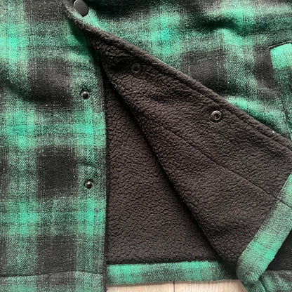 The Trapstar Irongate Green Plaid Lumberjack Jacket is a woven jacket in a green and black checked pattern, made from a 95% polyester and 5% wool outer with a full polyester lining. It features contrast shell panels at the shoulders, exposed popper fastenings at the front, a classic collar, and adjustable popper tabs at the cuffs. Details include side slip pockets, a Trapstar brand patch at the front hem, and a large Trapstar logo embroidered across the upper back yoke.