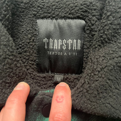 The Trapstar Irongate Green Plaid Lumberjack Jacket is a woven jacket in a green and black checked pattern, made from a 95% polyester and 5% wool outer with a full polyester lining. It features contrast shell panels at the shoulders, exposed popper fastenings at the front, a classic collar, and adjustable popper tabs at the cuffs. Details include side slip pockets, a Trapstar brand patch at the front hem, and a large Trapstar logo embroidered across the upper back yoke.