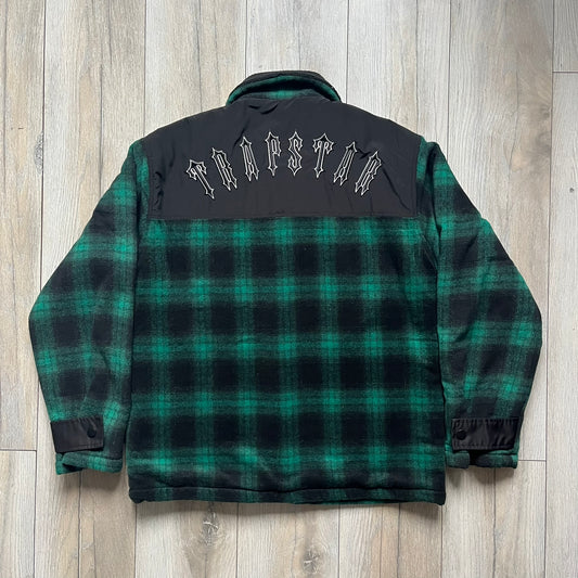 The Trapstar Irongate Green Plaid Lumberjack Jacket is a woven jacket in a green and black checked pattern, made from a 95% polyester and 5% wool outer with a full polyester lining. It features contrast shell panels at the shoulders, exposed popper fastenings at the front, a classic collar, and adjustable popper tabs at the cuffs. Details include side slip pockets, a Trapstar brand patch at the front hem, and a large Trapstar logo embroidered across the upper back yoke.