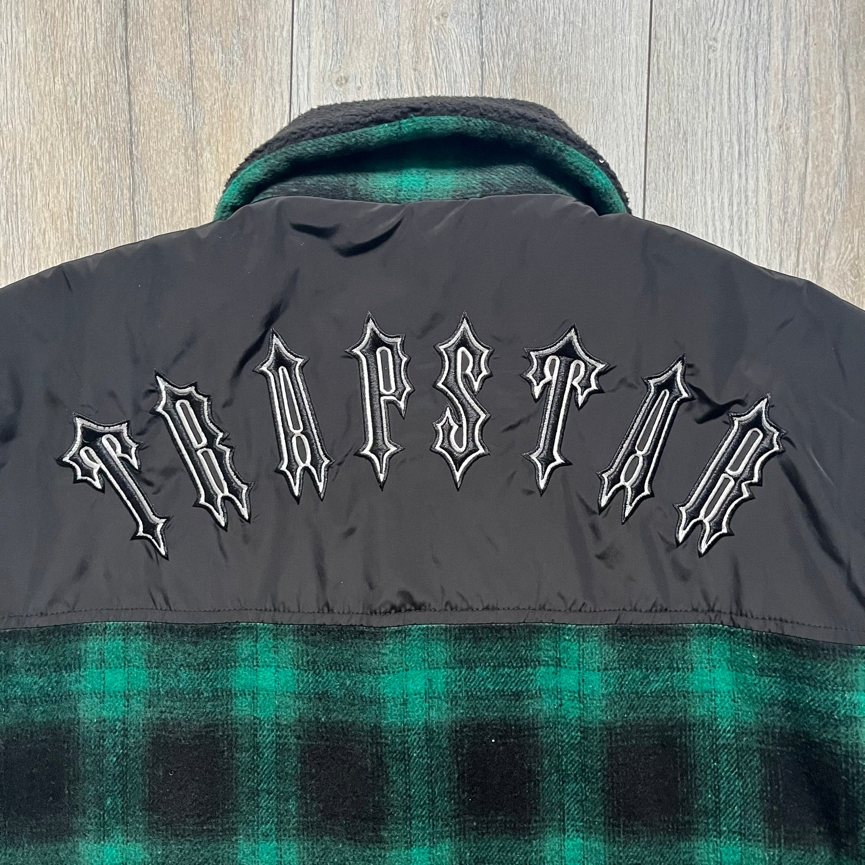The Trapstar Irongate Green Plaid Lumberjack Jacket is a woven jacket in a green and black checked pattern, made from a 95% polyester and 5% wool outer with a full polyester lining. It features contrast shell panels at the shoulders, exposed popper fastenings at the front, a classic collar, and adjustable popper tabs at the cuffs. Details include side slip pockets, a Trapstar brand patch at the front hem, and a large Trapstar logo embroidered across the upper back yoke.