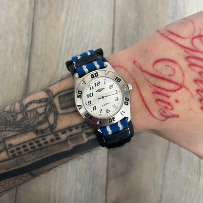 Umbro U528 The Double Diamond Analog Watch features a white dial with navy Umbro logo and numerals, silver and navy hands, and a silver case. The watch comes with a black Velcro strap accented with large white Umbro text and a small blue-and-white detail. It combines sporty design with a straightforward analog display.
