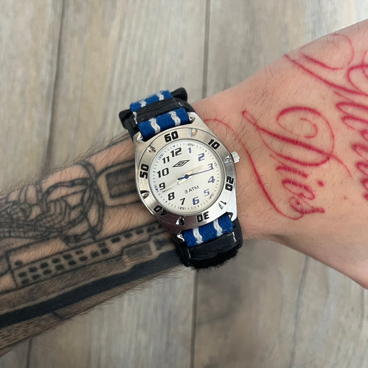 Umbro U528 The Double Diamond Analog Watch features a white dial with navy Umbro logo and numerals, silver and navy hands, and a silver case. The watch comes with a black Velcro strap accented with large white Umbro text and a small blue-and-white detail. It combines sporty design with a straightforward analog display.
