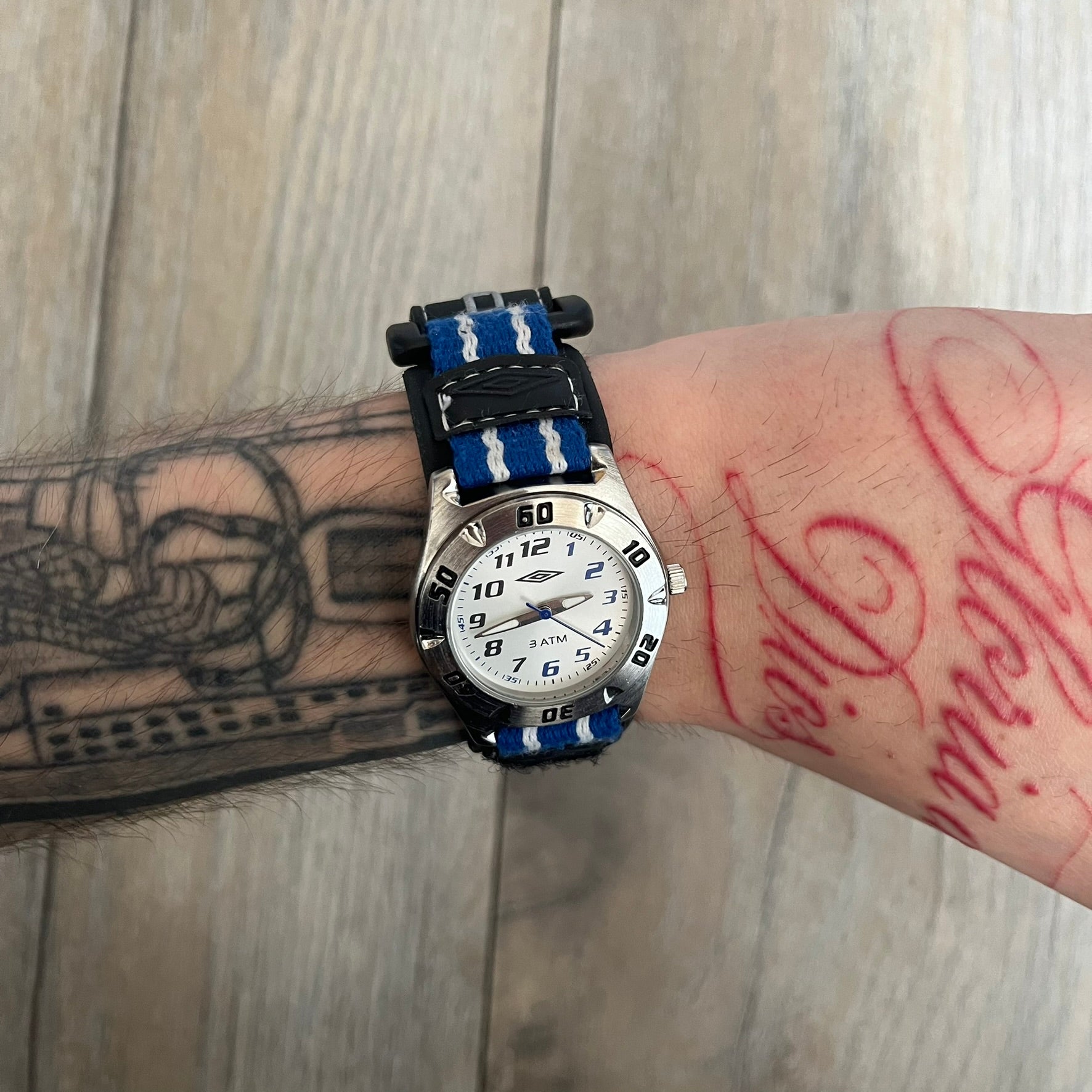 Umbro U528 The Double Diamond Analog Watch features a white dial with navy Umbro logo and numerals, silver and navy hands, and a silver case. The watch comes with a black Velcro strap accented with large white Umbro text and a small blue-and-white detail. It combines sporty design with a straightforward analog display.
