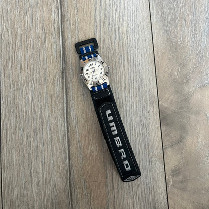 Umbro U528 The Double Diamond Analog Watch features a white dial with navy Umbro logo and numerals, silver and navy hands, and a silver case. The watch comes with a black Velcro strap accented with large white Umbro text and a small blue-and-white detail. It combines sporty design with a straightforward analog display.
