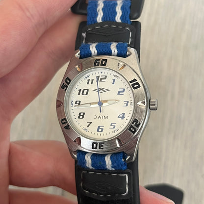 Umbro U528 The Double Diamond Analog Watch features a white dial with navy Umbro logo and numerals, silver and navy hands, and a silver case. The watch comes with a black Velcro strap accented with large white Umbro text and a small blue-and-white detail. It combines sporty design with a straightforward analog display.
