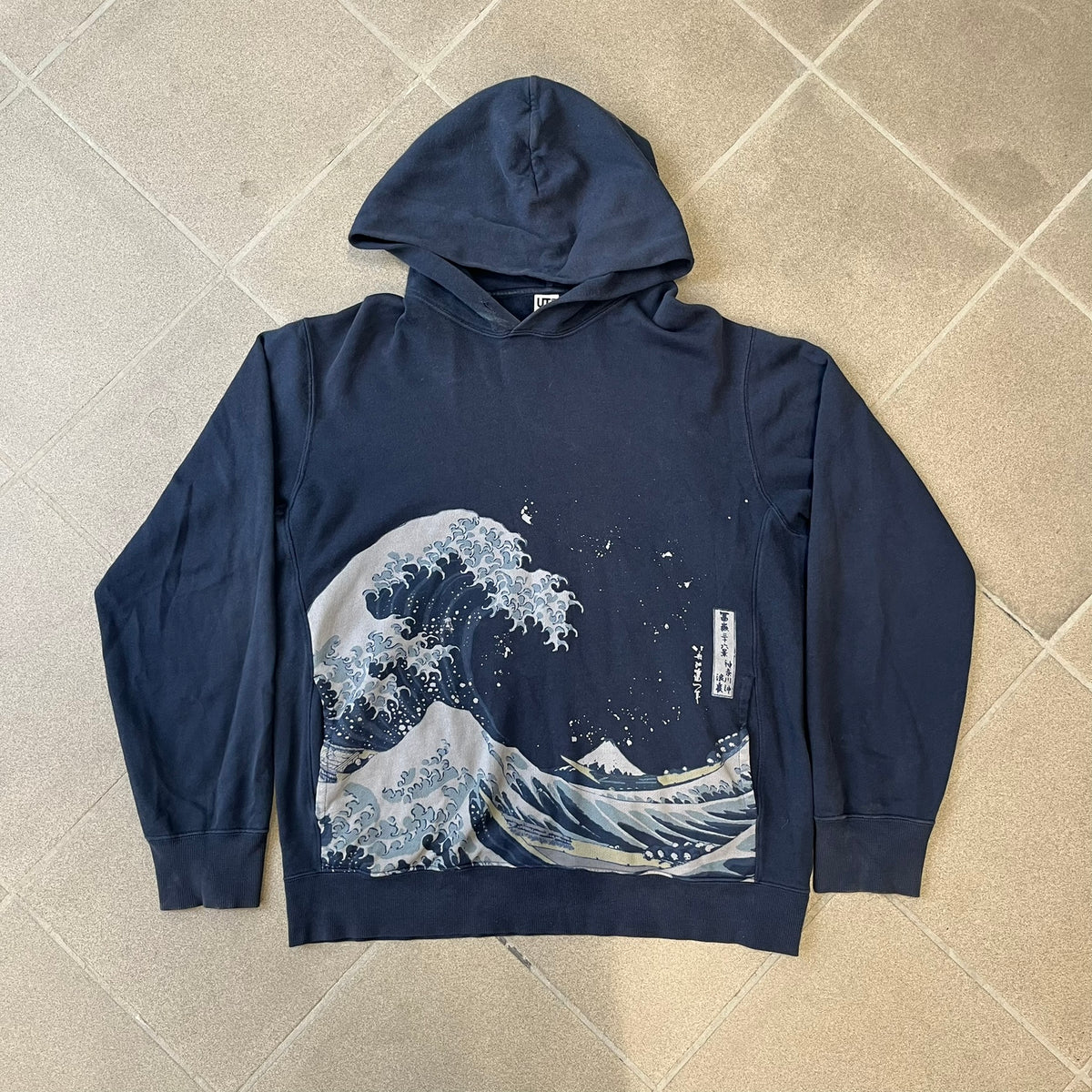 Uniqlo UT 2018 Hokusai The Great Wave Hoodie is a thick, dark navy hoodie featuring a large print of "The Great Wave off Kanagawa" by Hokusai across the lower front, created in collaboration with the Museum of Fine Arts Boston. It includes side pockets.