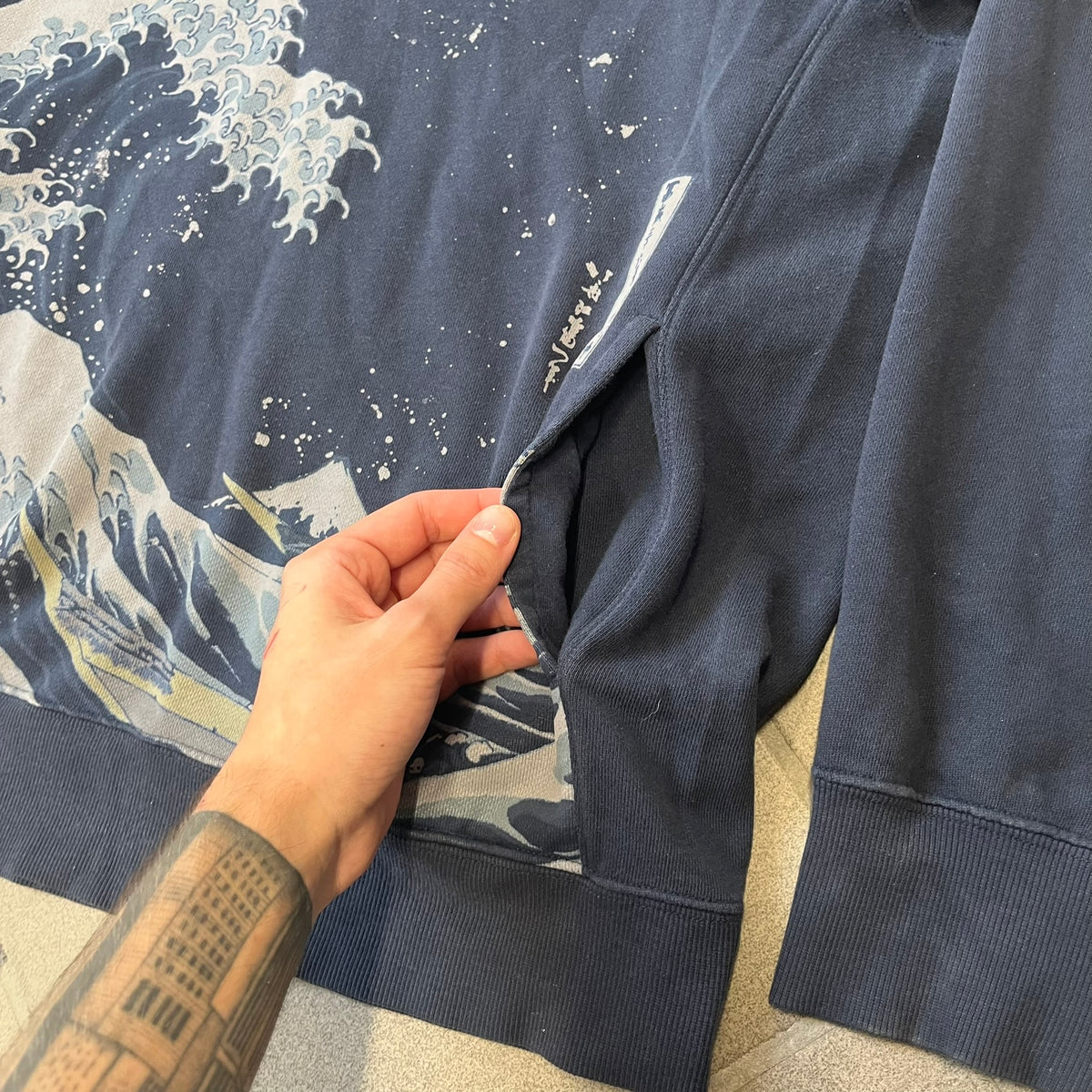 Uniqlo UT 2018 Hokusai The Great Wave Hoodie is a thick, dark navy hoodie featuring a large print of "The Great Wave off Kanagawa" by Hokusai across the lower front, created in collaboration with the Museum of Fine Arts Boston. It includes side pockets.