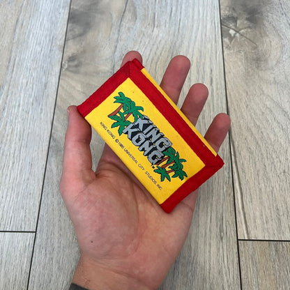 Universal Studios Florida 1985 King Kong Key Holder Wallet is a yellow wallet with red trim featuring King Kong-themed graphics on both sides. One side displays a King Kong illustration with Universal Studios Florida branding, while the other features the King Kong logo, palm tree graphics, and 1985 licensing details. The interior includes a storage pocket and four metal key holders for organizing keys.
