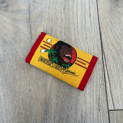 Universal Studios Florida 1985 King Kong Key Holder Wallet is a yellow wallet with red trim featuring King Kong-themed graphics on both sides. One side displays a King Kong illustration with Universal Studios Florida branding, while the other features the King Kong logo, palm tree graphics, and 1985 licensing details. The interior includes a storage pocket and four metal key holders for organizing keys.
