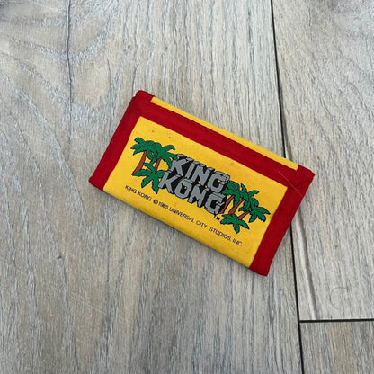 Universal Studios Florida 1985 King Kong Key Holder Wallet is a yellow wallet with red trim featuring King Kong-themed graphics on both sides. One side displays a King Kong illustration with Universal Studios Florida branding, while the other features the King Kong logo, palm tree graphics, and 1985 licensing details. The interior includes a storage pocket and four metal key holders for organizing keys.
