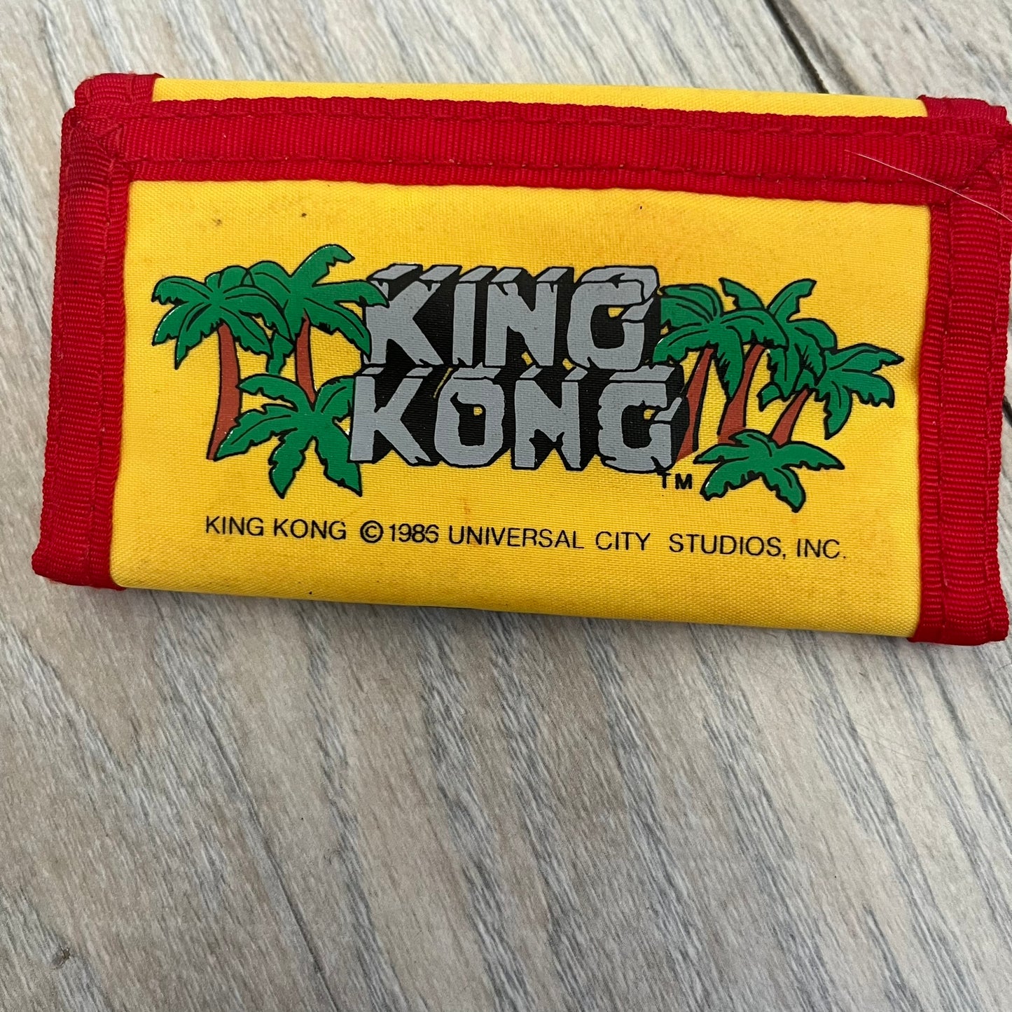 Universal Studios Florida 1985 King Kong Key Holder Wallet is a yellow wallet with red trim featuring King Kong-themed graphics on both sides. One side displays a King Kong illustration with Universal Studios Florida branding, while the other features the King Kong logo, palm tree graphics, and 1985 licensing details. The interior includes a storage pocket and four metal key holders for organizing keys.
