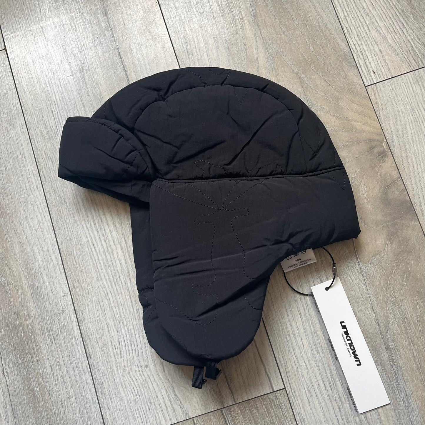 Unknown London Dagger Stitch Trapper Hat is a black trapper-style hat made from nylon and polyester. It features detailed tonal stitching with patterned dagger-style motifs, earflaps, and an adjustable clasp closure at the bottom. The hat comes with the original dust bag.