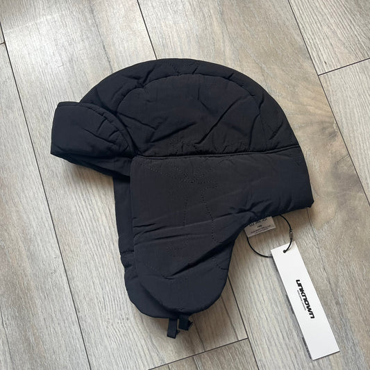 Unknown London Dagger Stitch Trapper Hat is a black trapper-style hat made from nylon and polyester. It features detailed tonal stitching with patterned dagger-style motifs, earflaps, and an adjustable clasp closure at the bottom. The hat comes with the original dust bag.