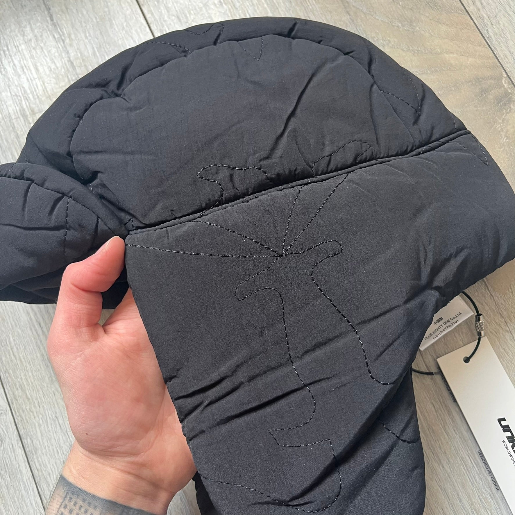 Unknown London Dagger Stitch Trapper Hat is a black trapper-style hat made from nylon and polyester. It features detailed tonal stitching with patterned dagger-style motifs, earflaps, and an adjustable clasp closure at the bottom. The hat comes with the original dust bag.