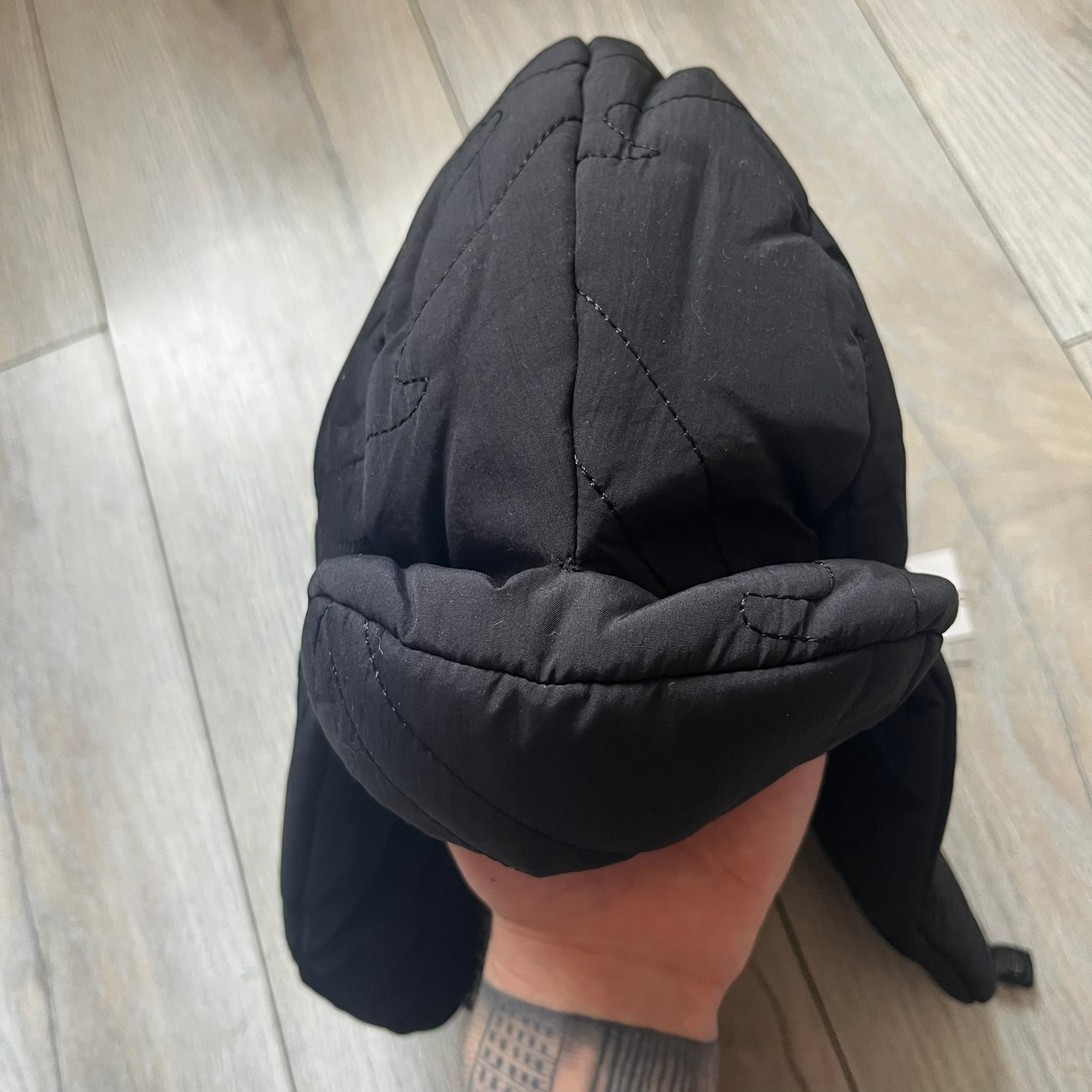 Unknown London Dagger Stitch Trapper Hat is a black trapper-style hat made from nylon and polyester. It features detailed tonal stitching with patterned dagger-style motifs, earflaps, and an adjustable clasp closure at the bottom. The hat comes with the original dust bag.