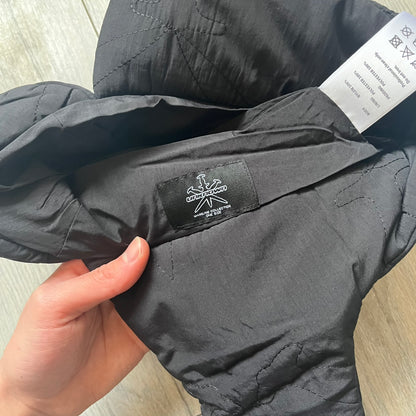 Unknown London Dagger Stitch Trapper Hat is a black trapper-style hat made from nylon and polyester. It features detailed tonal stitching with patterned dagger-style motifs, earflaps, and an adjustable clasp closure at the bottom. The hat comes with the original dust bag.