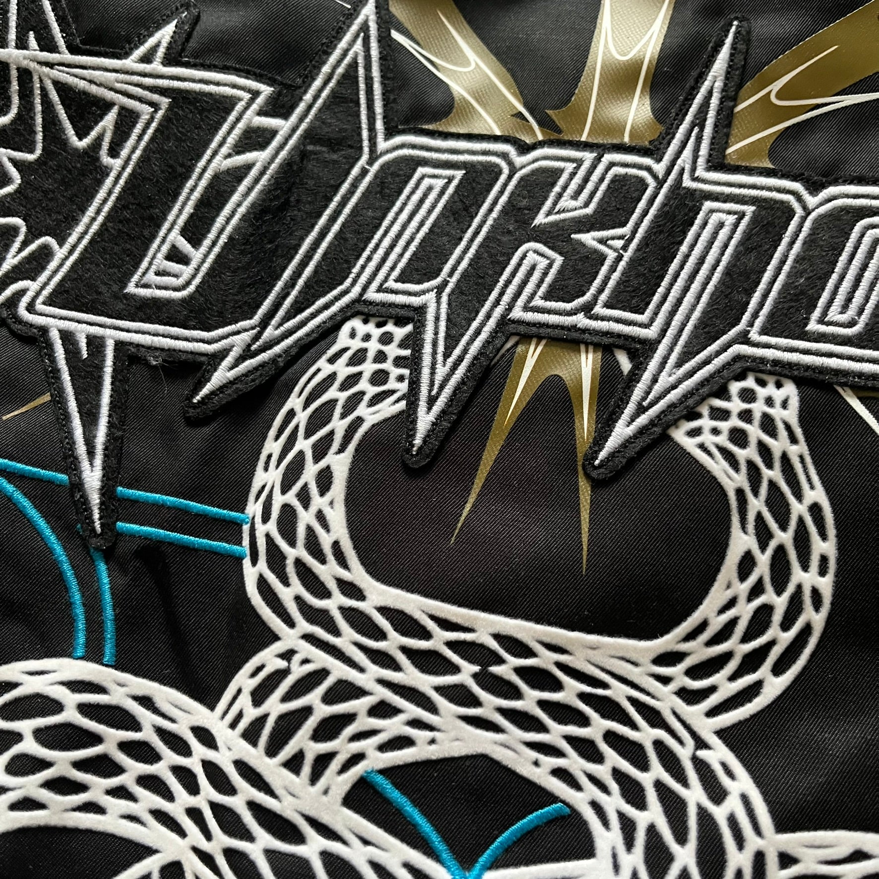 Unknown London Reversible Bomber Jacket crafted from padded polyester with a relaxed fit. One side features black matte fabric with embroidered logo, snakes, and multicolor detailing, plus arm patch and chest logo. The reverse side is beige with subtle chest embroidery in a softer cotton-like texture, offering two distinct styling options.