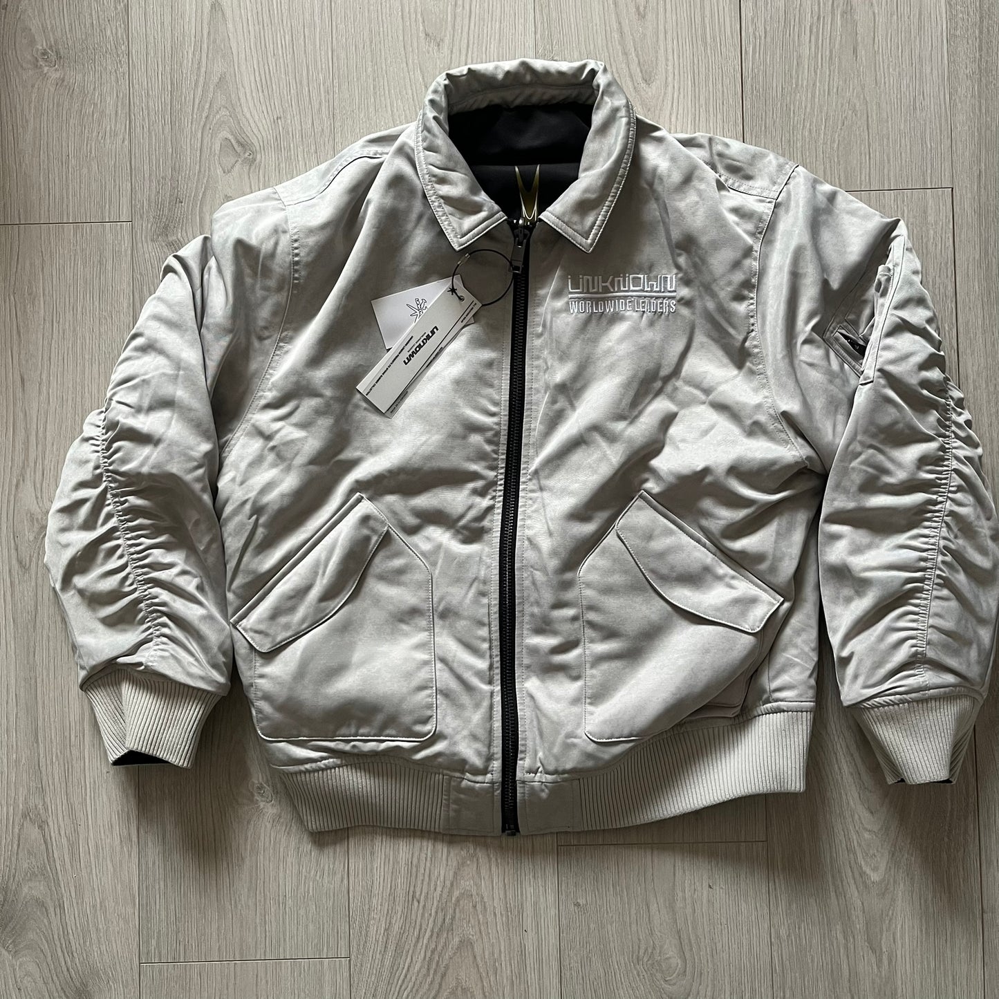 Unknown London Reversible Bomber Jacket crafted from padded polyester with a relaxed fit. One side features black matte fabric with embroidered logo, snakes, and multicolor detailing, plus arm patch and chest logo. The reverse side is beige with subtle chest embroidery in a softer cotton-like texture, offering two distinct styling options.