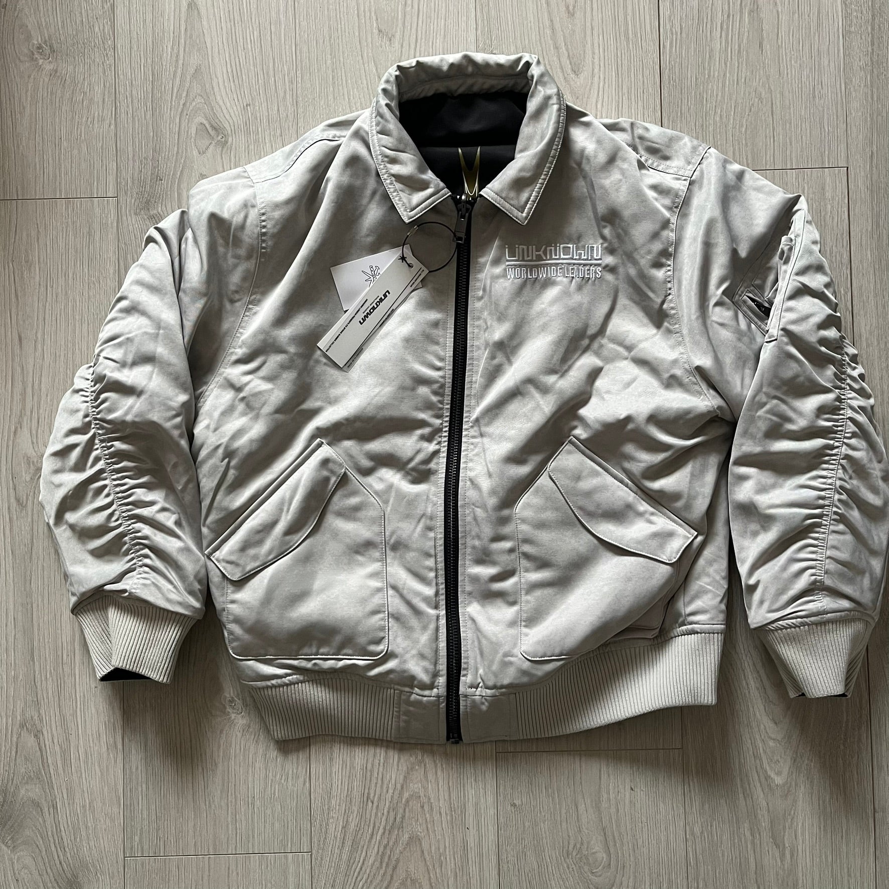 Unknown London Reversible Bomber Jacket crafted from padded polyester with a relaxed fit. One side features black matte fabric with embroidered logo, snakes, and multicolor detailing, plus arm patch and chest logo. The reverse side is beige with subtle chest embroidery in a softer cotton-like texture, offering two distinct styling options.