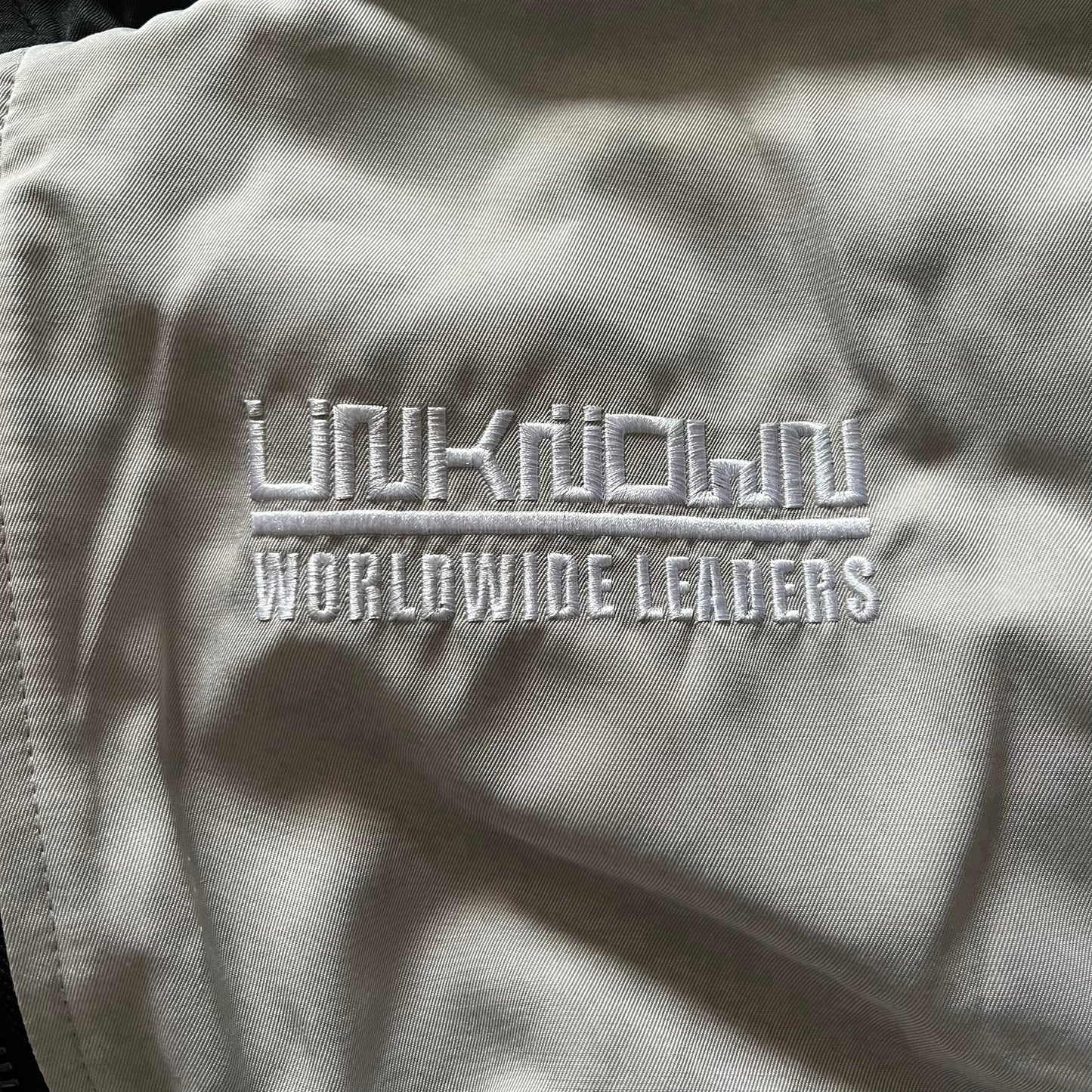 Unknown London Reversible Bomber Jacket crafted from padded polyester with a relaxed fit. One side features black matte fabric with embroidered logo, snakes, and multicolor detailing, plus arm patch and chest logo. The reverse side is beige with subtle chest embroidery in a softer cotton-like texture, offering two distinct styling options.