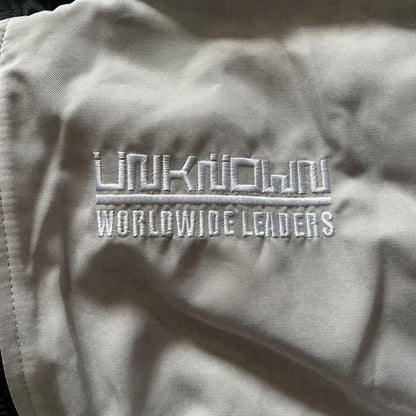 Unknown London Reversible Bomber Jacket crafted from padded polyester with a relaxed fit. One side features black matte fabric with embroidered logo, snakes, and multicolor detailing, plus arm patch and chest logo. The reverse side is beige with subtle chest embroidery in a softer cotton-like texture, offering two distinct styling options.