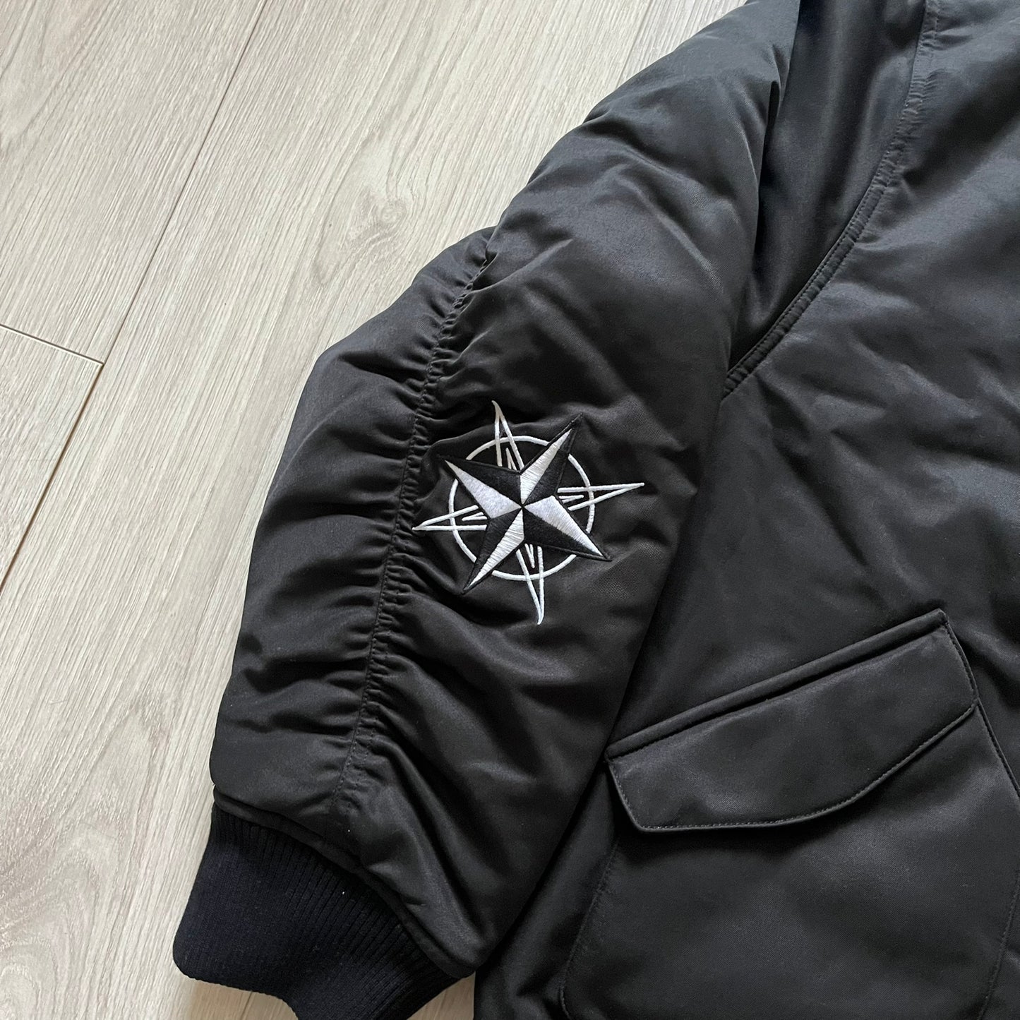 Unknown London Reversible Bomber Jacket crafted from padded polyester with a relaxed fit. One side features black matte fabric with embroidered logo, snakes, and multicolor detailing, plus arm patch and chest logo. The reverse side is beige with subtle chest embroidery in a softer cotton-like texture, offering two distinct styling options.