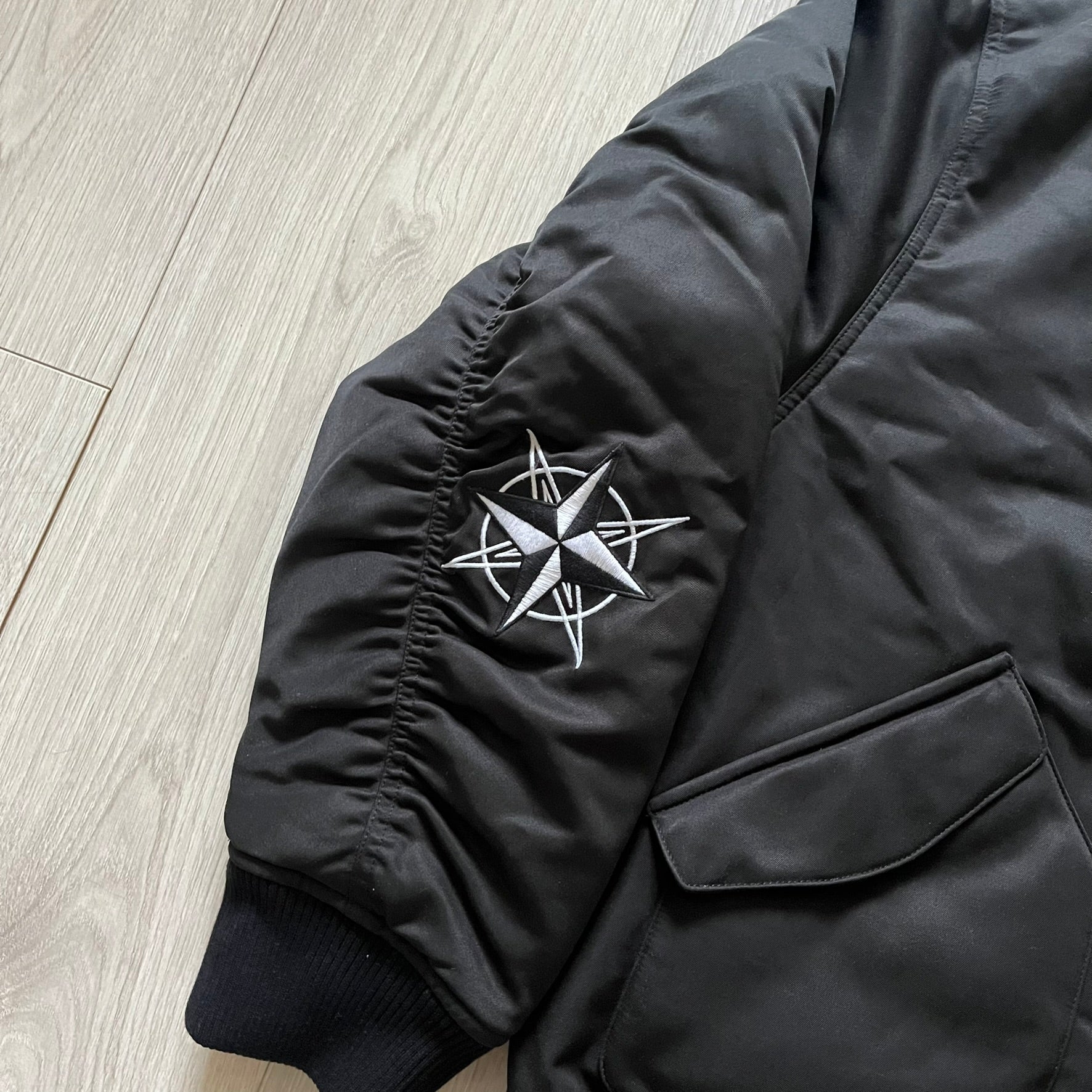 Unknown London Reversible Bomber Jacket crafted from padded polyester with a relaxed fit. One side features black matte fabric with embroidered logo, snakes, and multicolor detailing, plus arm patch and chest logo. The reverse side is beige with subtle chest embroidery in a softer cotton-like texture, offering two distinct styling options.