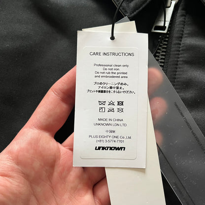 Unknown London Reversible Bomber Jacket crafted from padded polyester with a relaxed fit. One side features black matte fabric with embroidered logo, snakes, and multicolor detailing, plus arm patch and chest logo. The reverse side is beige with subtle chest embroidery in a softer cotton-like texture, offering two distinct styling options.