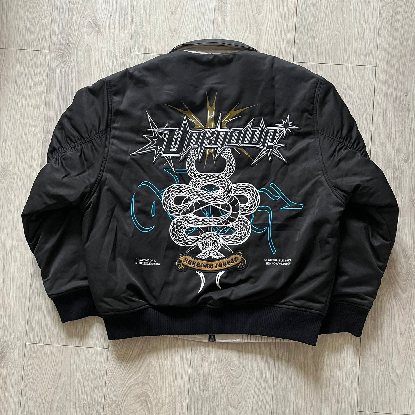 Unknown London Reversible Bomber Jacket crafted from padded polyester with a relaxed fit. One side features black matte fabric with embroidered logo, snakes, and multicolor detailing, plus arm patch and chest logo. The reverse side is beige with subtle chest embroidery in a softer cotton-like texture, offering two distinct styling options.