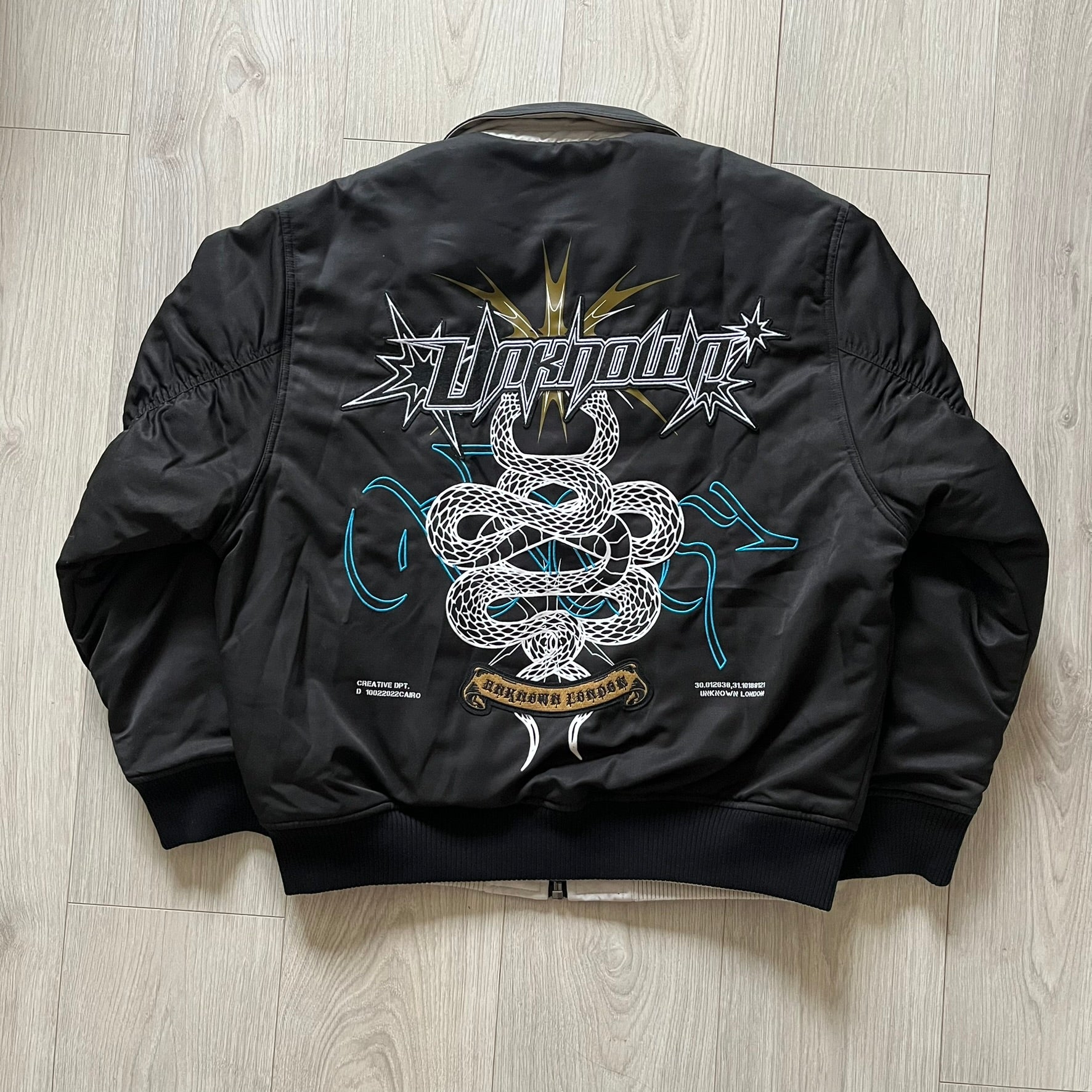 Unknown London Reversible Bomber Jacket crafted from padded polyester with a relaxed fit. One side features black matte fabric with embroidered logo, snakes, and multicolor detailing, plus arm patch and chest logo. The reverse side is beige with subtle chest embroidery in a softer cotton-like texture, offering two distinct styling options.