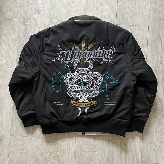 Unknown London Reversible Bomber Jacket crafted from padded polyester with a relaxed fit. One side features black matte fabric with embroidered logo, snakes, and multicolor detailing, plus arm patch and chest logo. The reverse side is beige with subtle chest embroidery in a softer cotton-like texture, offering two distinct styling options.