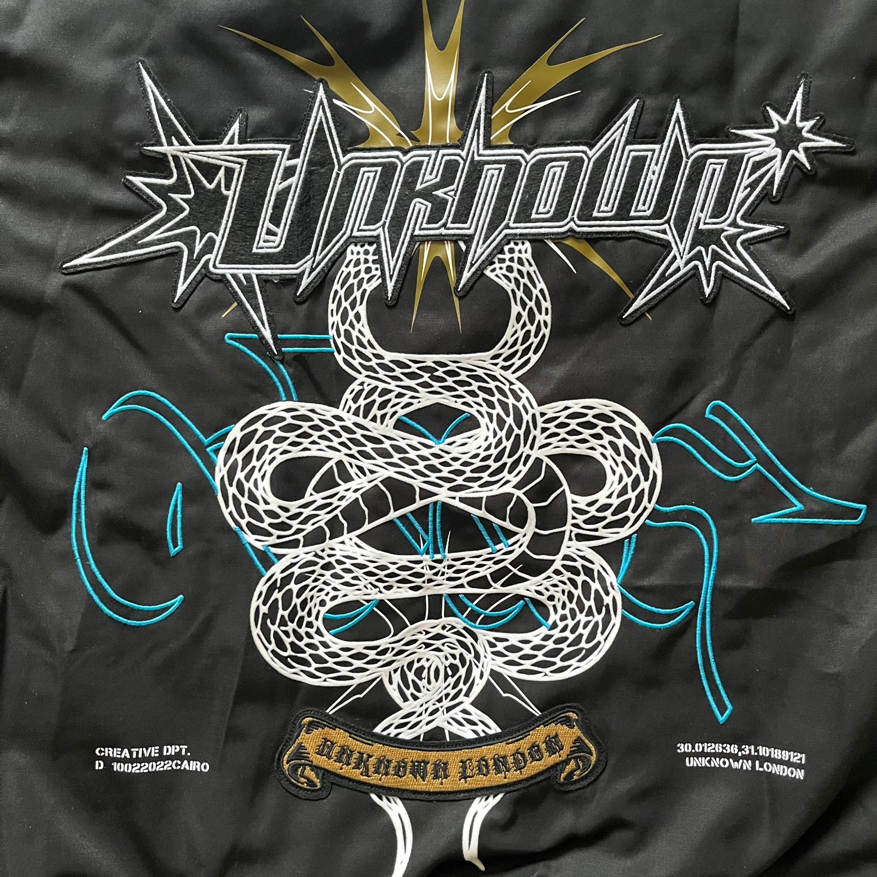 Unknown London Reversible Bomber Jacket crafted from padded polyester with a relaxed fit. One side features black matte fabric with embroidered logo, snakes, and multicolor detailing, plus arm patch and chest logo. The reverse side is beige with subtle chest embroidery in a softer cotton-like texture, offering two distinct styling options.