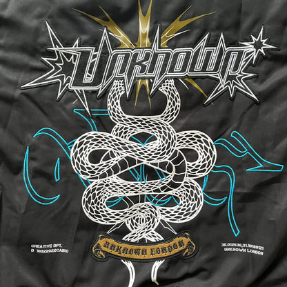 Unknown London Reversible Bomber Jacket crafted from padded polyester with a relaxed fit. One side features black matte fabric with embroidered logo, snakes, and multicolor detailing, plus arm patch and chest logo. The reverse side is beige with subtle chest embroidery in a softer cotton-like texture, offering two distinct styling options.