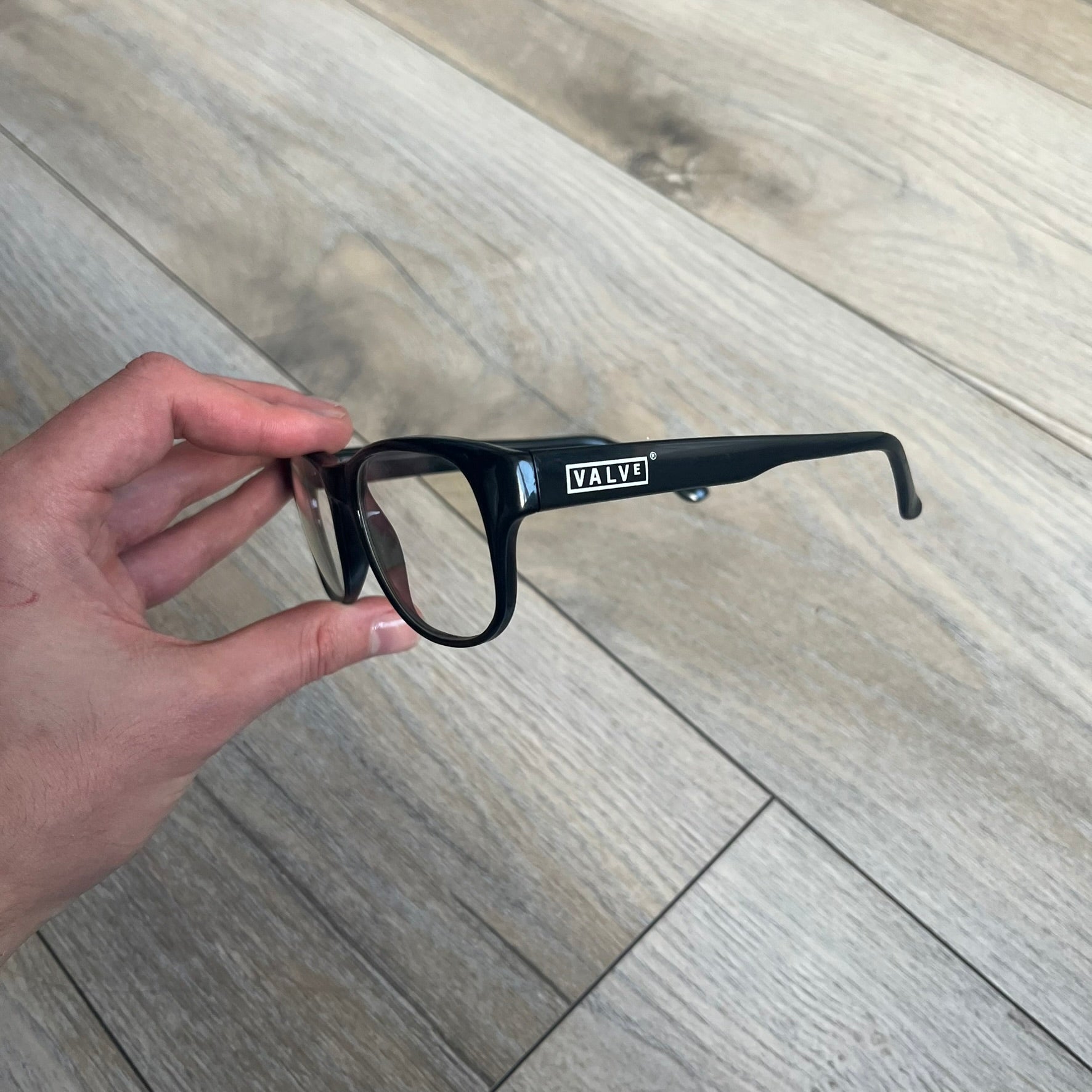 Valve Black Promo Eyeglasses are non-prescription promotional glasses featuring simple black plastic frames with clear zero-strength lenses. Small white Valve logos are placed on both sides of the frame arms. The design is minimal and intended as branded promotional eyewear.