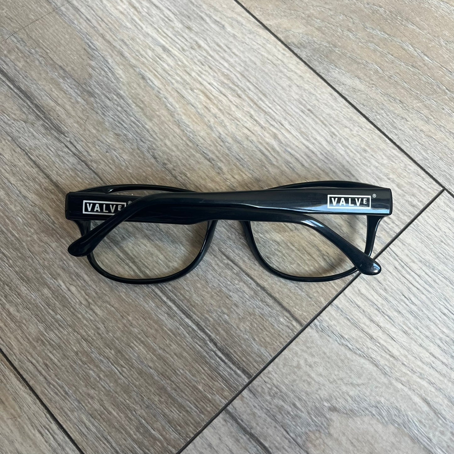 Valve Black Promo Eyeglasses are non-prescription promotional glasses featuring simple black plastic frames with clear zero-strength lenses. Small white Valve logos are placed on both sides of the frame arms. The design is minimal and intended as branded promotional eyewear.