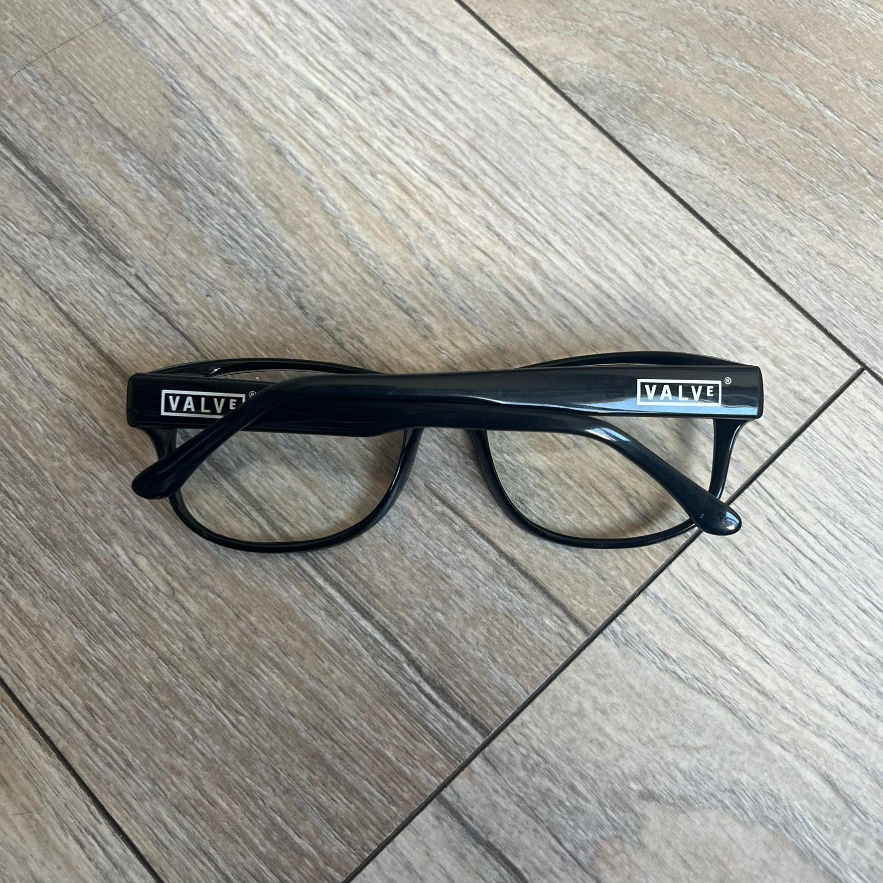 Valve Black Promo Eyeglasses are non-prescription promotional glasses featuring simple black plastic frames with clear zero-strength lenses. Small white Valve logos are placed on both sides of the frame arms. The design is minimal and intended as branded promotional eyewear.