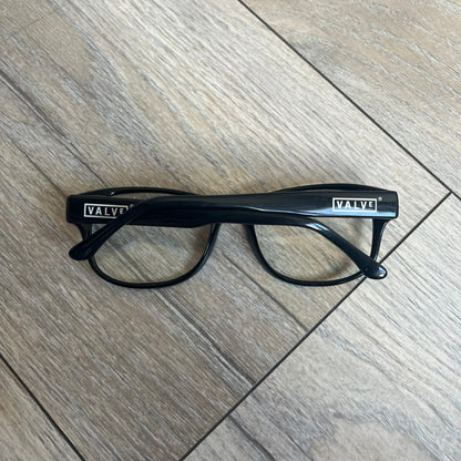 Valve Black Promo Eyeglasses are non-prescription promotional glasses featuring simple black plastic frames with clear zero-strength lenses. Small white Valve logos are placed on both sides of the frame arms. The design is minimal and intended as branded promotional eyewear.