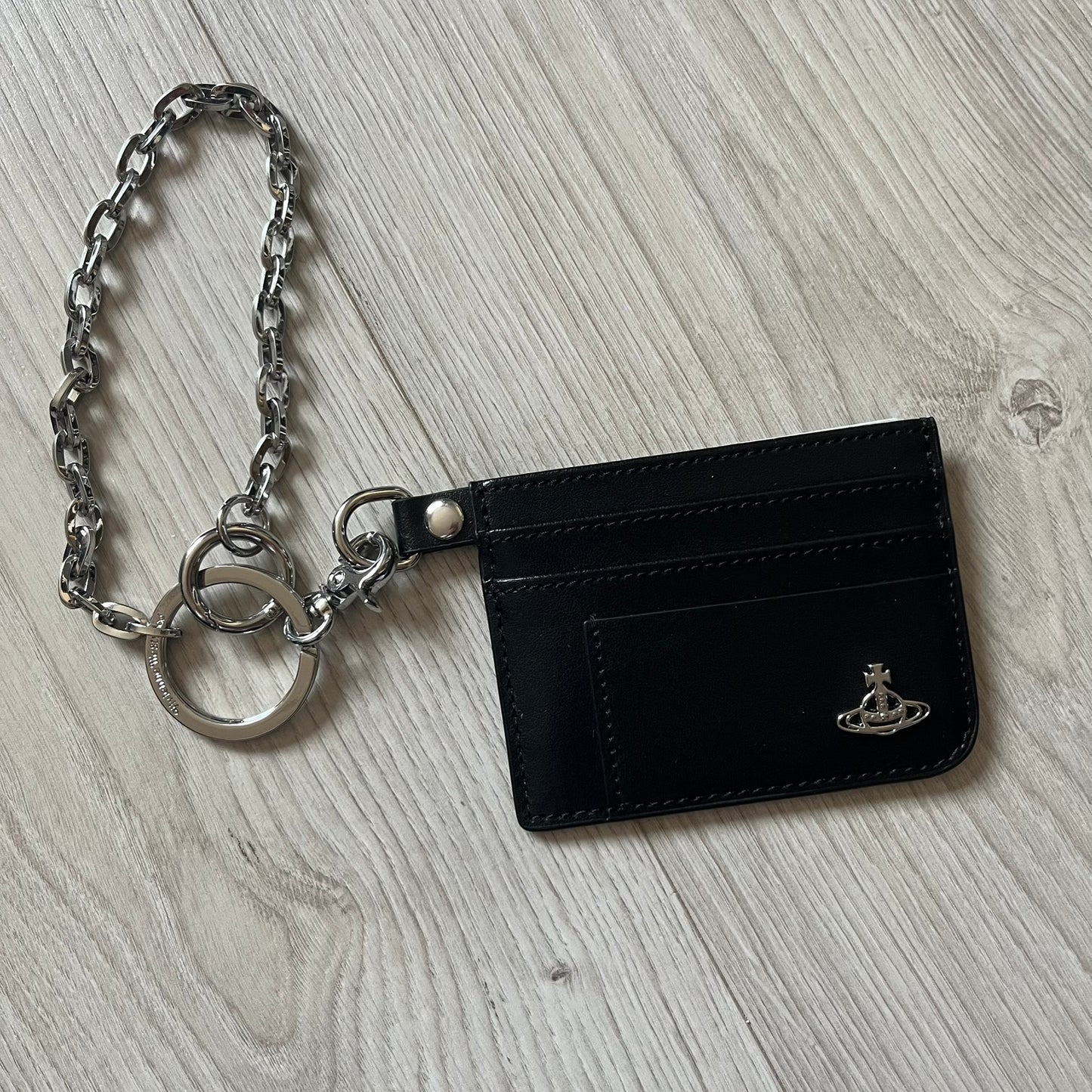 Vivienne Westwood Flat Chain Card Holder in black leather, featuring the signature silver Orb emblem on the corner. Designed with four compartments - three main slots and one smaller front pocket. Completed with a flat chain and an engraved Vivienne Westwood loop for a refined finish.