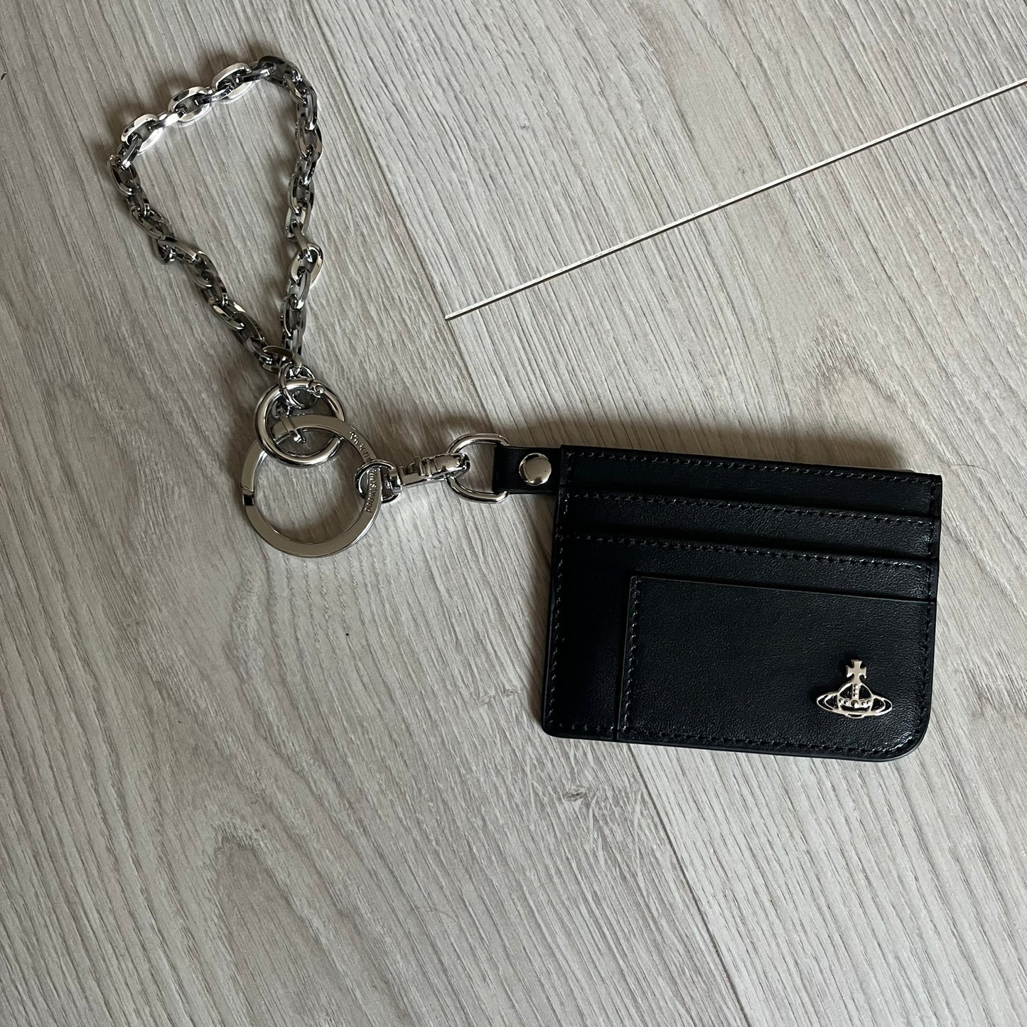 Vivienne Westwood Flat Chain Card Holder in black leather, featuring the signature silver Orb emblem on the corner. Designed with four compartments - three main slots and one smaller front pocket. Completed with a flat chain and an engraved Vivienne Westwood loop for a refined finish.