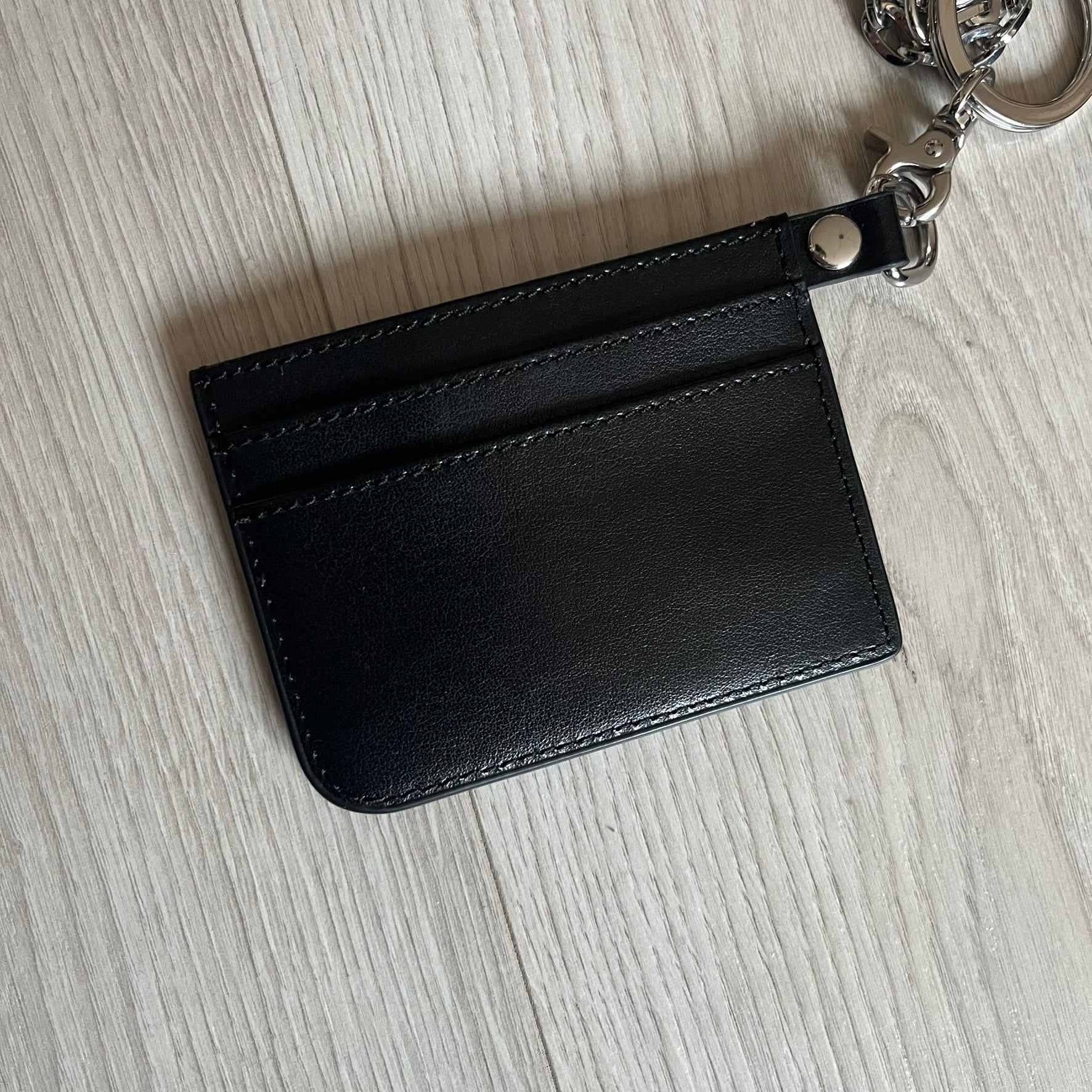Vivienne Westwood Flat Chain Card Holder in black leather, featuring the signature silver Orb emblem on the corner. Designed with four compartments - three main slots and one smaller front pocket. Completed with a flat chain and an engraved Vivienne Westwood loop for a refined finish.
