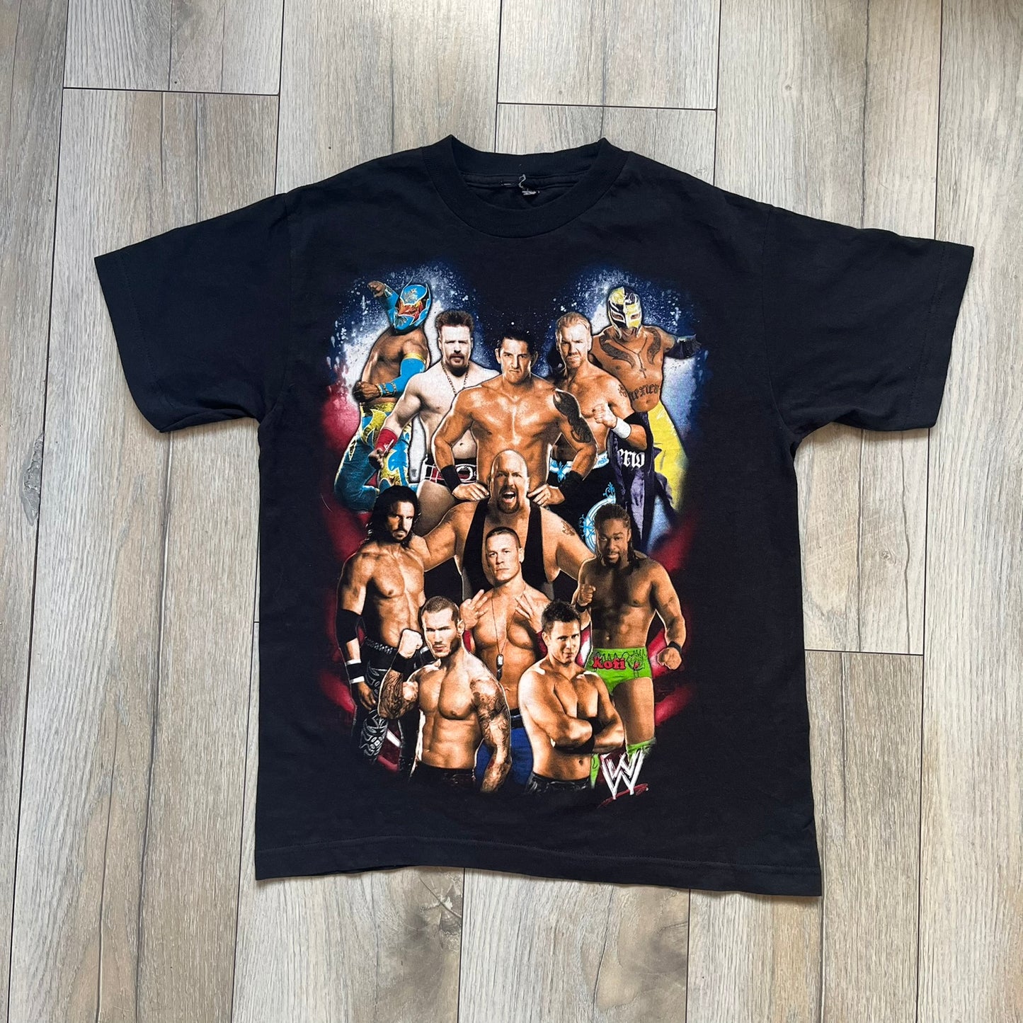 WWE 2011 All Over Print Promo Tee is a black short-sleeve T-shirt featuring a large front graphic of multiple WWE wrestlers, including Rey Mysterio, John Cena, and Randy Orton. A small WWE logo is printed beneath the main graphic. The shirt has no neck tag and was released as official promotional merchandise in 2011.