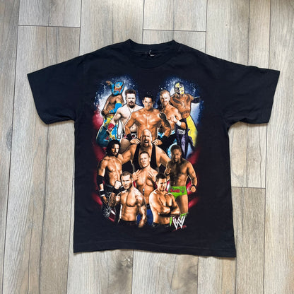 WWE 2011 All Over Print Promo Tee is a black short-sleeve T-shirt featuring a large front graphic of multiple WWE wrestlers, including Rey Mysterio, John Cena, and Randy Orton. A small WWE logo is printed beneath the main graphic. The shirt has no neck tag and was released as official promotional merchandise in 2011.