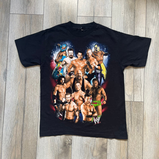 WWE 2011 All Over Print Promo Tee is a black short-sleeve T-shirt featuring a large front graphic of multiple WWE wrestlers, including Rey Mysterio, John Cena, and Randy Orton. A small WWE logo is printed beneath the main graphic. The shirt has no neck tag and was released as official promotional merchandise in 2011.