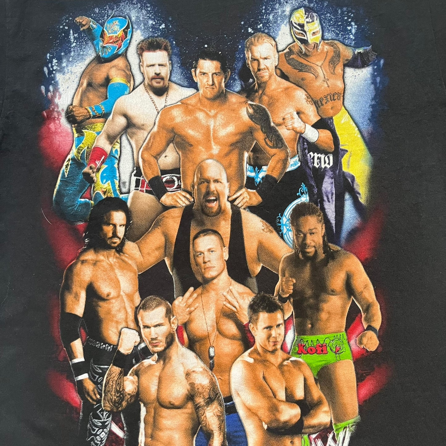 WWE 2011 All Over Print Promo Tee is a black short-sleeve T-shirt featuring a large front graphic of multiple WWE wrestlers, including Rey Mysterio, John Cena, and Randy Orton. A small WWE logo is printed beneath the main graphic. The shirt has no neck tag and was released as official promotional merchandise in 2011.