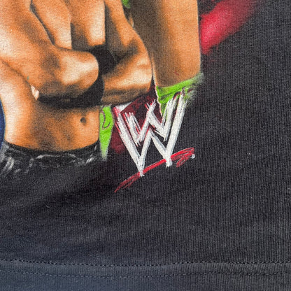 WWE 2011 All Over Print Promo Tee is a black short-sleeve T-shirt featuring a large front graphic of multiple WWE wrestlers, including Rey Mysterio, John Cena, and Randy Orton. A small WWE logo is printed beneath the main graphic. The shirt has no neck tag and was released as official promotional merchandise in 2011.