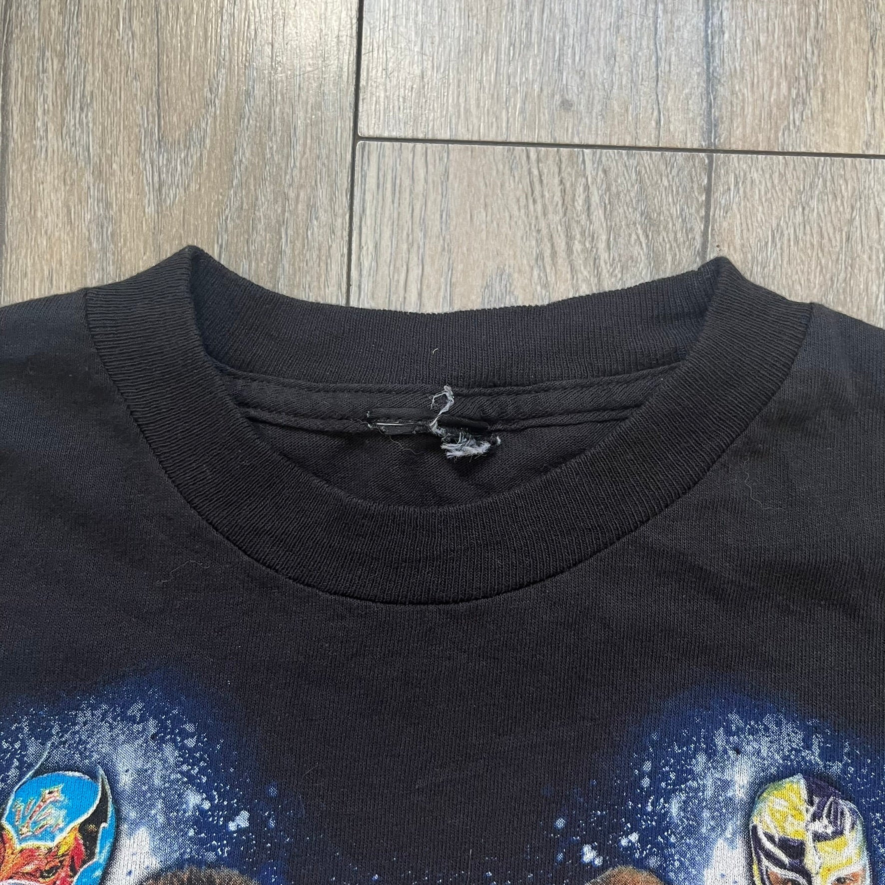 WWE 2011 All Over Print Promo Tee is a black short-sleeve T-shirt featuring a large front graphic of multiple WWE wrestlers, including Rey Mysterio, John Cena, and Randy Orton. A small WWE logo is printed beneath the main graphic. The shirt has no neck tag and was released as official promotional merchandise in 2011.