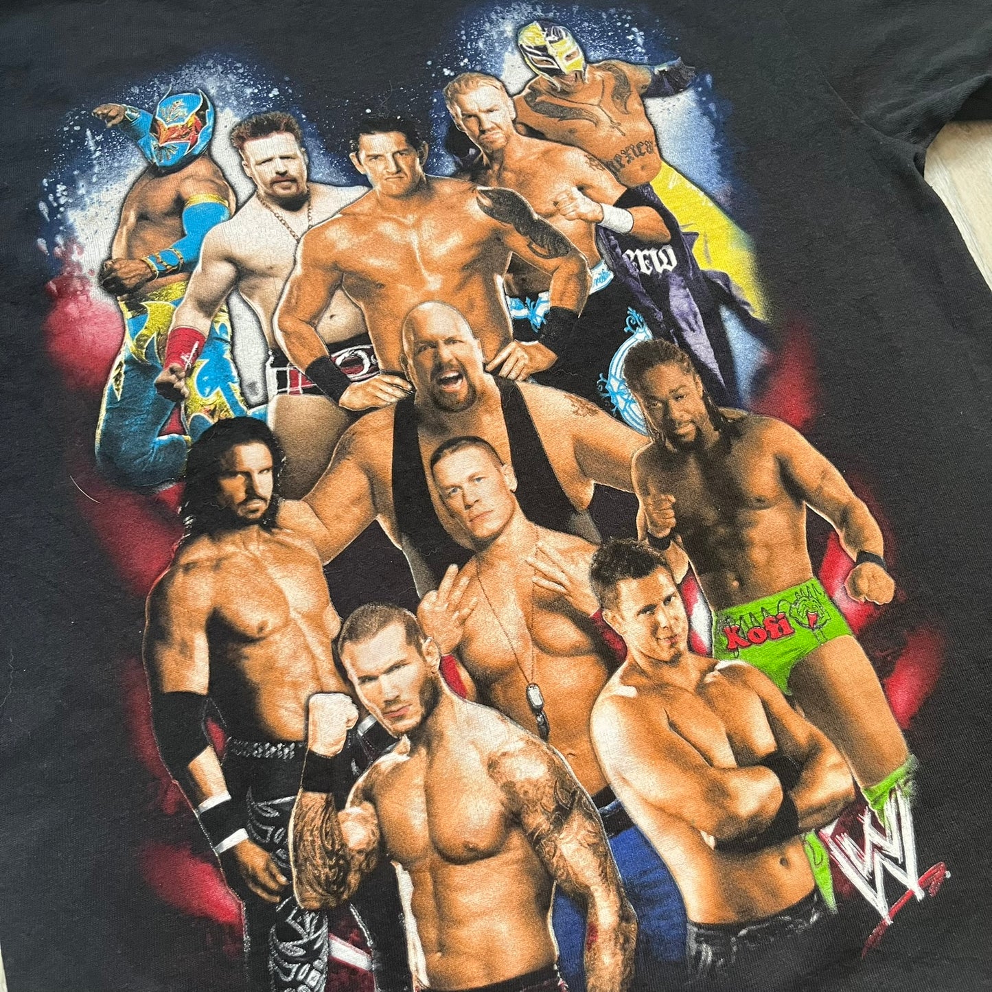 WWE 2011 All Over Print Promo Tee is a black short-sleeve T-shirt featuring a large front graphic of multiple WWE wrestlers, including Rey Mysterio, John Cena, and Randy Orton. A small WWE logo is printed beneath the main graphic. The shirt has no neck tag and was released as official promotional merchandise in 2011.