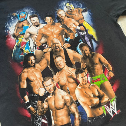 WWE 2011 All Over Print Promo Tee is a black short-sleeve T-shirt featuring a large front graphic of multiple WWE wrestlers, including Rey Mysterio, John Cena, and Randy Orton. A small WWE logo is printed beneath the main graphic. The shirt has no neck tag and was released as official promotional merchandise in 2011.